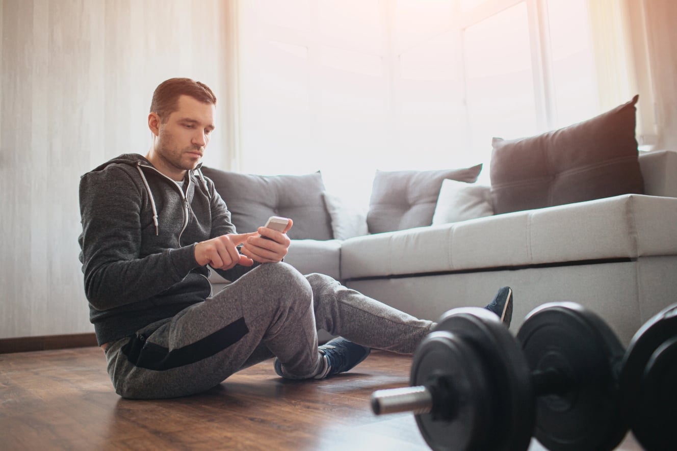 Uomo seduto davanti ai pesi che guarda lo smartphone – tipica distrazione durante l’allenamento a casa