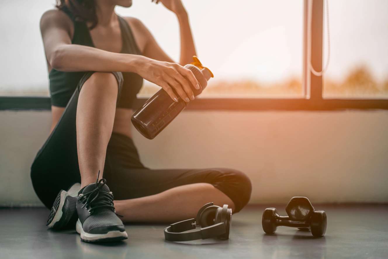 Donna in palestra che si rilassa dopo l'allenamento con bottiglia d'acqua, manubri e cuffie in primo piano.