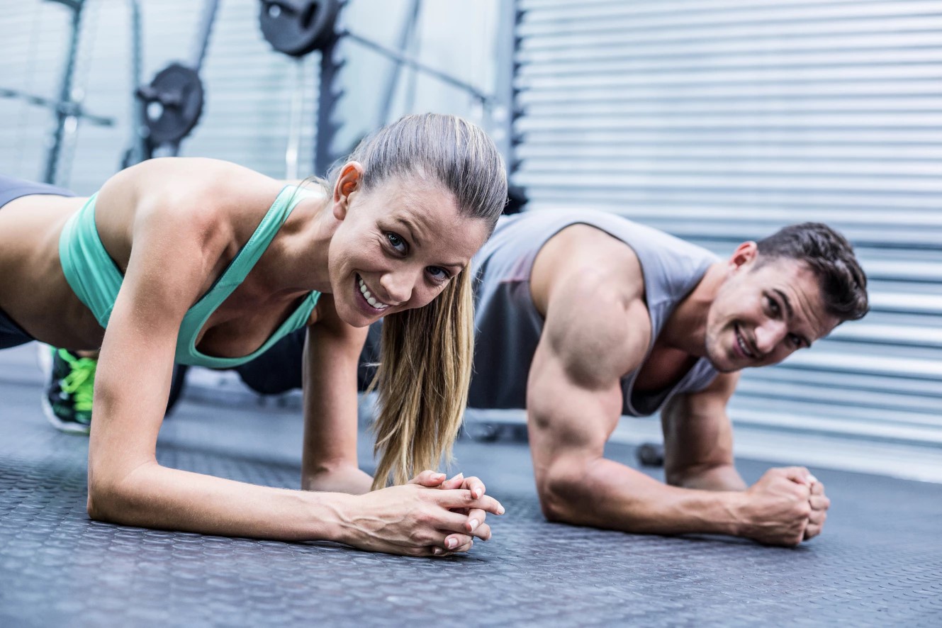 Un uomo e una donna in una palestra sorridono e fanno un esercizio di supporto per gli avambracci su un tappetino grigio.